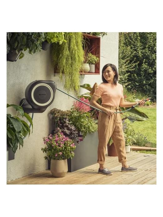 גלגלת צינור אוטומטית GARDENA מותקנת על קיר במרפסת עם עציצים, אישה משקה בצינור - משתלות הדר נוי