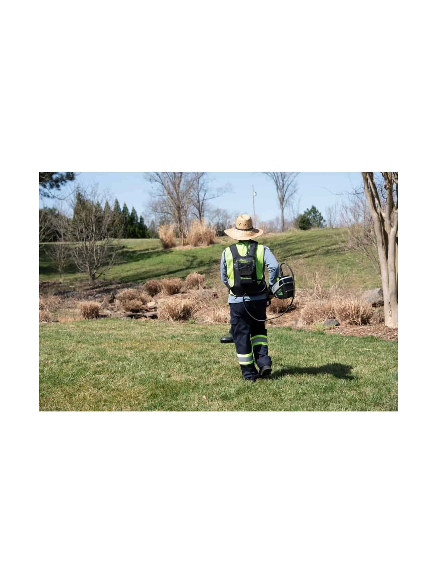 חרמש 41 ס״מ (16 אינץ׳) 80 וולט נטען, Greenworks STB460: המפעיל מגבו הולך על מדשאה, עם רתמת הסוללה על הגב והחרמש ביד