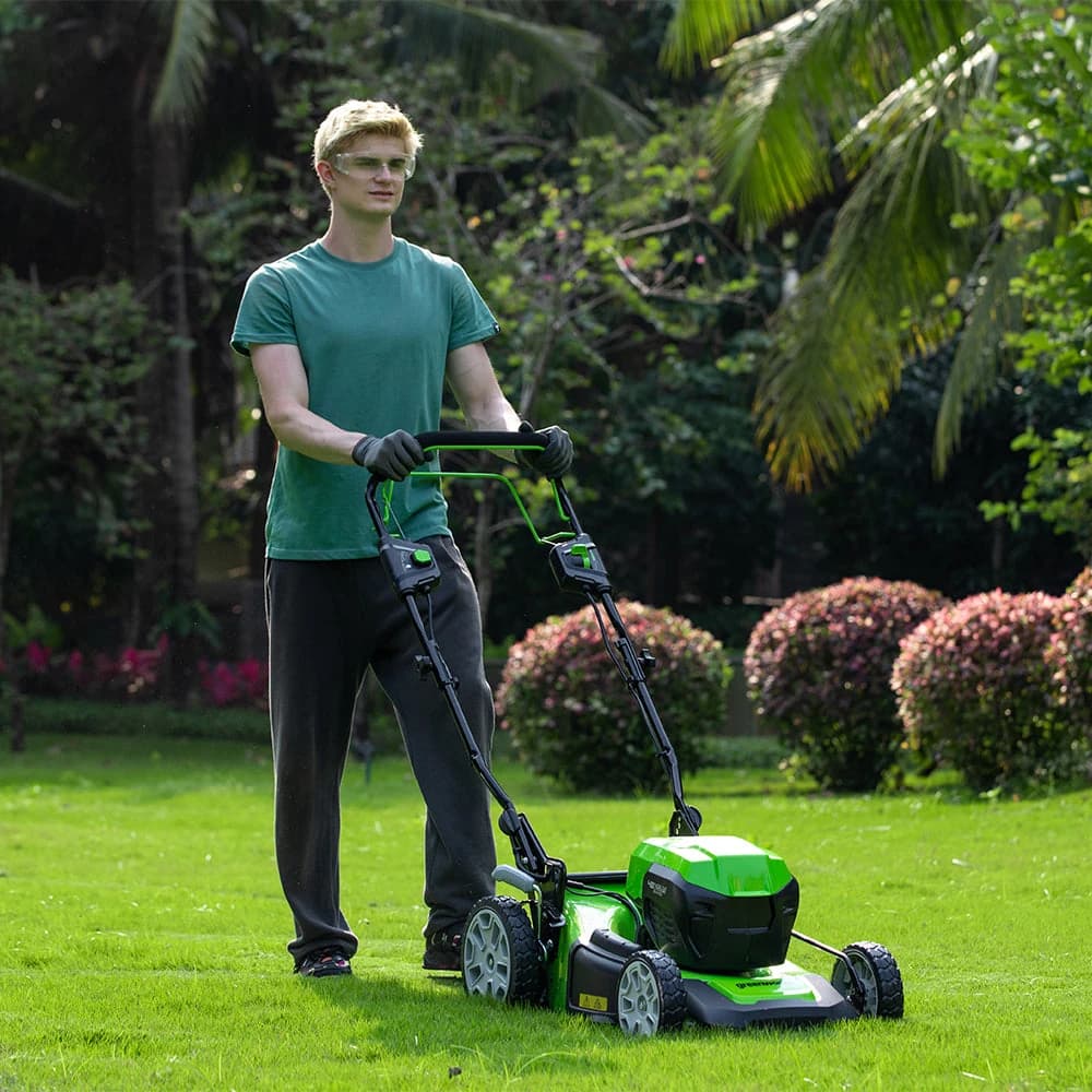 מכסחת דשא 46 ס״מ 48 וולט נטענת נסיעה עצמית, Greenworks GD24X2LM46SP: כיסוח מדשאה בגינה עם שיחים פורחים, המפעיל מול המצלמה