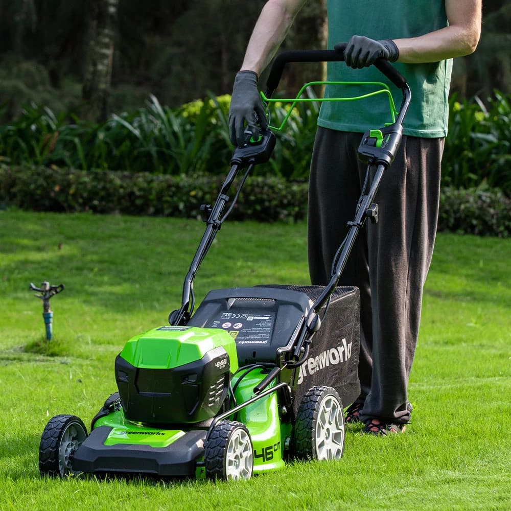 מכסחת דשא 46 ס״מ 48 וולט נטענת נסיעה עצמית, Greenworks GD24X2LM46SP: המכסחת מתקדמת על הדשא מול המצלמה, עם ממטרה ברקע