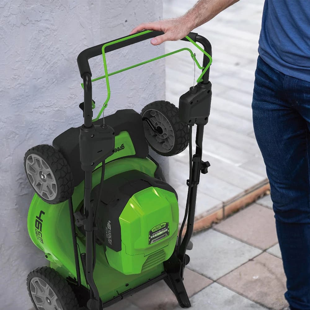 מכסחת דשא 46 ס״מ 48 וולט נטענת נסיעה עצמית, Greenworks GD24X2LM46SP: המכסחת מקופלת ועומדת זקופה ליד קיר, מוחזקת ביד אחת