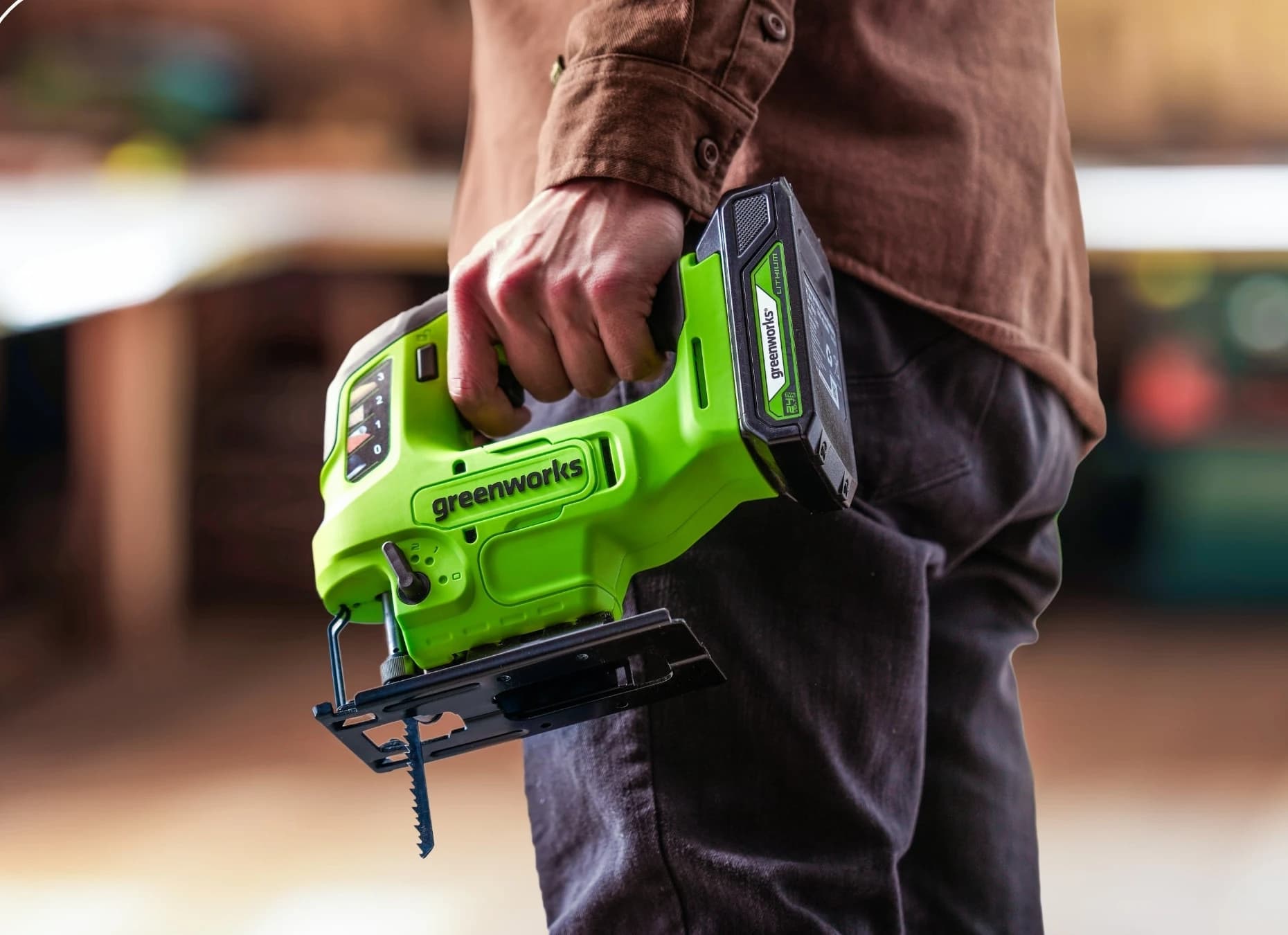 מסור אנכי 24 וולט נטען דור 2, Greenworks JS24B340: המסור עם סוללה מחוברת נישא ביד לצד הגוף בסדנה, בלי כיתוב