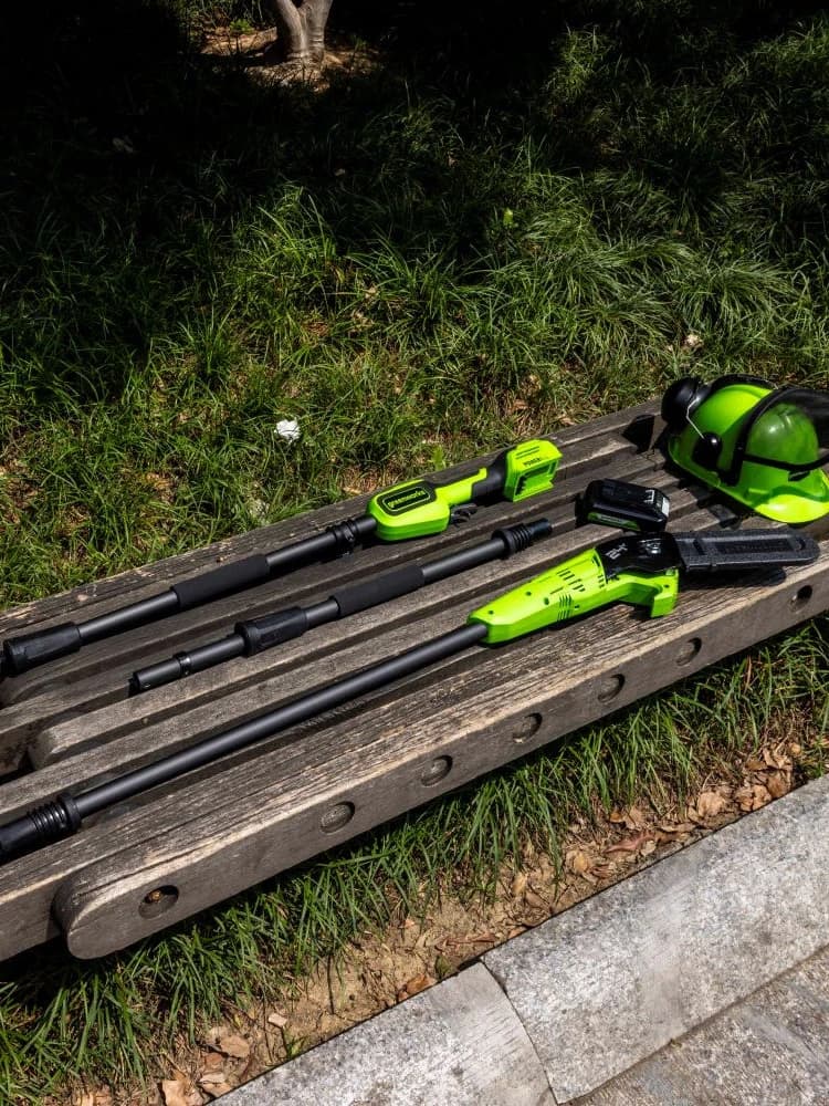 מסור גבהים 20 ס״מ 24 וולט נטען דור 2 גוף בלבד, Greenworks PS24B00-01: חלקי הכלי פרוסים על ספסל עץ בגינה: שלושה חלקי מוט, ראש המסור, סוללה וקסדת מגן