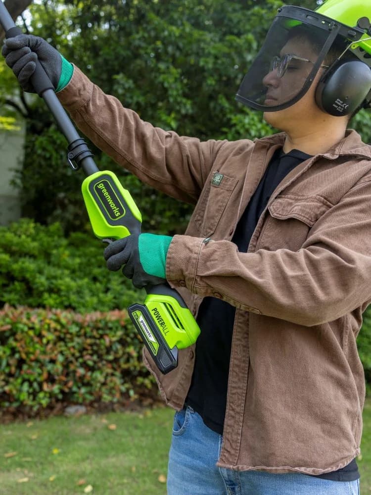 מסור גבהים 20 ס״מ 24 וולט נטען דור 2 גוף בלבד, Greenworks PS24B00-01: מבט מקרוב על האחיזה בשתי ידיים בזמן ניסור מעל הראש, עם קסדה ומגני אוזניים